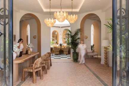 Grande salle de restaurant avec des banquettes et coussins en velours rouge, des chaises en rotin et des tables dressées. L'espace est décoré dans un style méditerranéen avec un sol en terre cuite, des arches murales ocre, et des suspensions en paille tressée