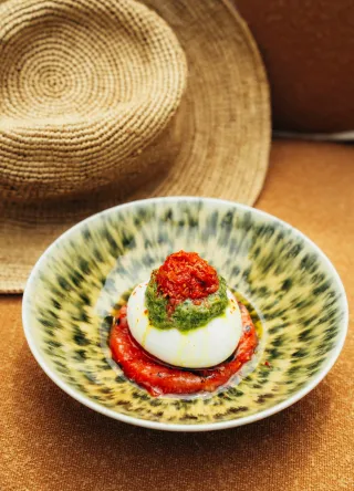 A nicely dressed dish containing a burrata or mozzarella dome on a bed of red tomato sauce, topped with a layer of green pesto and another dark red condiment, presented in a leopard/camouflage bowl, with a straw hat in the background