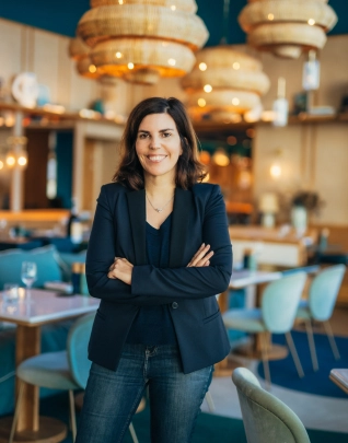 Empresaria sonriente con un blazer azul oscuro y jeans, brazos cruzados, de pie en un restaurante moderno y luminoso. El fondo borroso presenta mesas, sillas y lámparas colgantes altas de mimbre de color azul verdoso