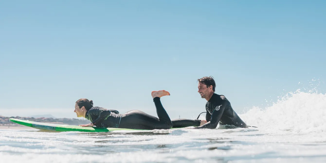 A man and a woman wearing wetsuits on surfboards, waiting for a wave in the ocean. The woman is lying on his board while the man is rowing. The sky is blue and clear