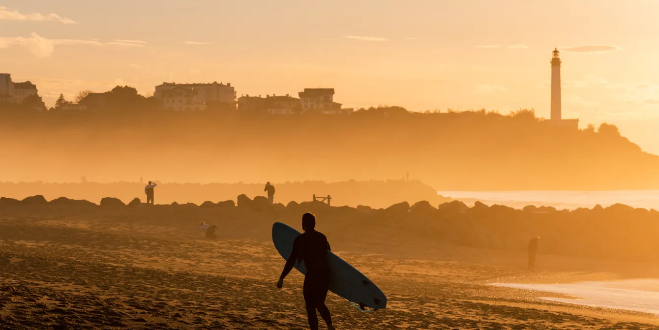Silhouette of a surfer carrying his board on a sandy beach at sunset. The background is bathed in a misty orange light, showing cliffs, buildings, and the Biarritz lighthouse in the distance