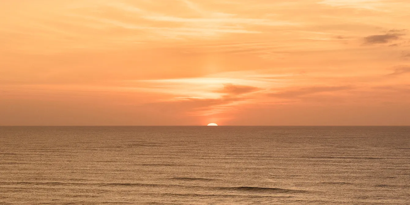 Ocean sunset, with the disc of the Sun disappearing just below the horizon, painting the sky in warm orange and gold hues above the still water