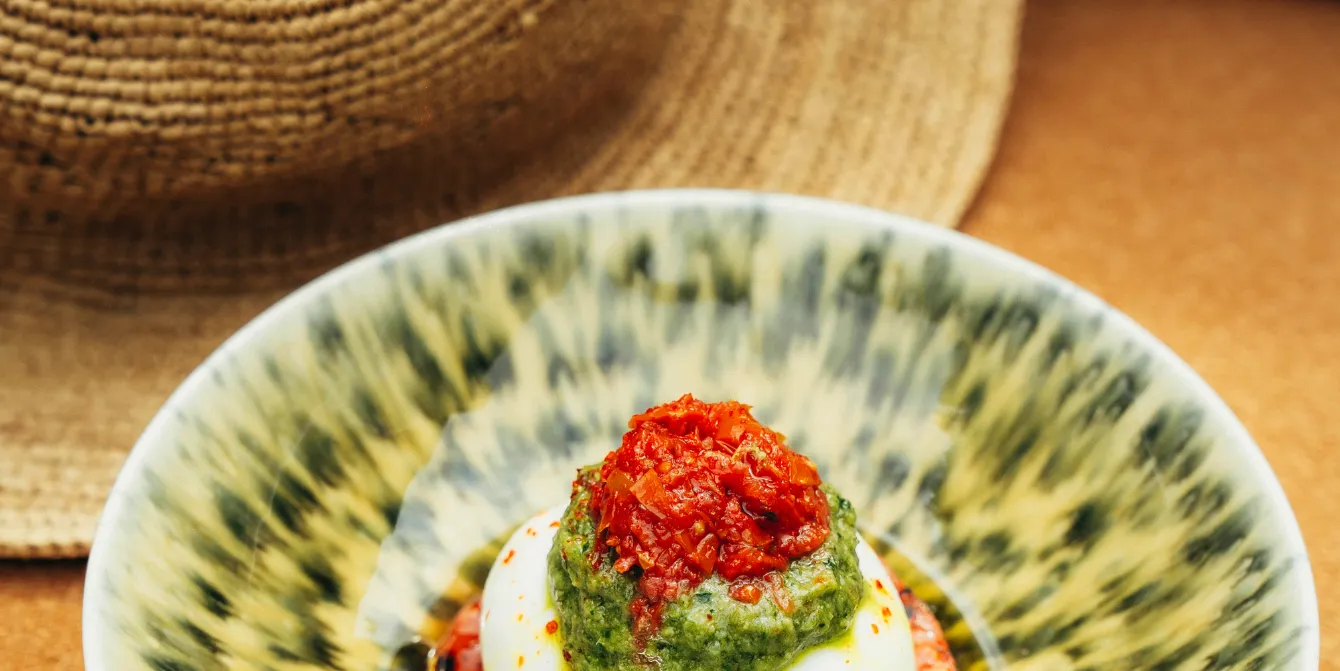Close-up on an Italian burrata or mozzarella dish topped with green sauce (pesto) and dried red tomato relish, served on a decorative green and yellow plate. A straw hat is visible in the background