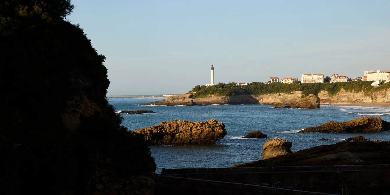 View of the rocky coastline of Biarritz at sunset, with the famous lighthouse in the distance at the top of the hill. Brown rocks are visible in the water in front of a steep coastline