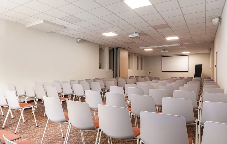 Amplia y moderna sala de conferencias o anfiteatro, amueblada en estilo teatro con numerosas hileras de sillas blancas y naranjas. La sala está equipada con una gran pantalla de proyección y un proyector de techo