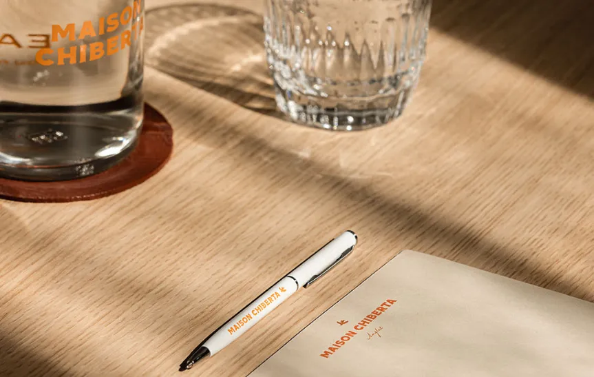 Acércate a un escritorio de madera clara bañado por la luz del sol, con un bolígrafo y un bloc de notas de la marca «Maison Chiberta». También vemos un vaso de agua y un posavasos de piel.