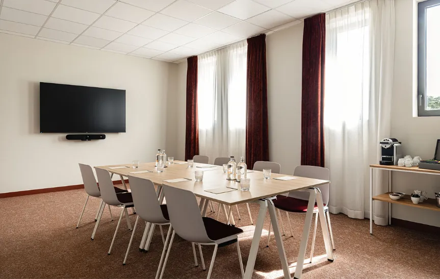 Moderna sala de reuniones con una gran mesa rectangular rodeada de sillas grises y burdeos, equipada con botellas de agua y vasos. La sala tiene una gran pantalla de televisión montada en la pared, ventanas con cortinas blancas y burdeos y una zona de café