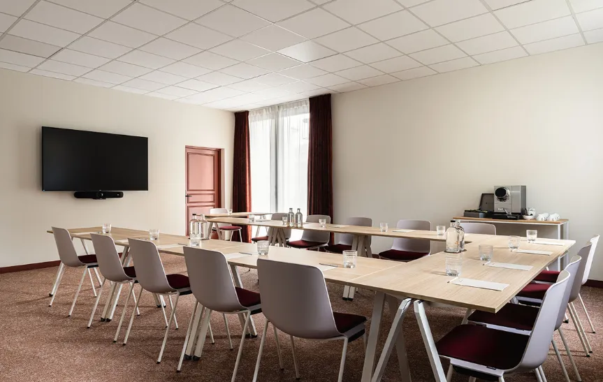 Sala de conferencias profesional con una gran mesa en forma de U, rodeada de sillas grises y burdeos. La sala está equipada con una pantalla grande, un televisor montado en la pared y una zona de café en el lateral.