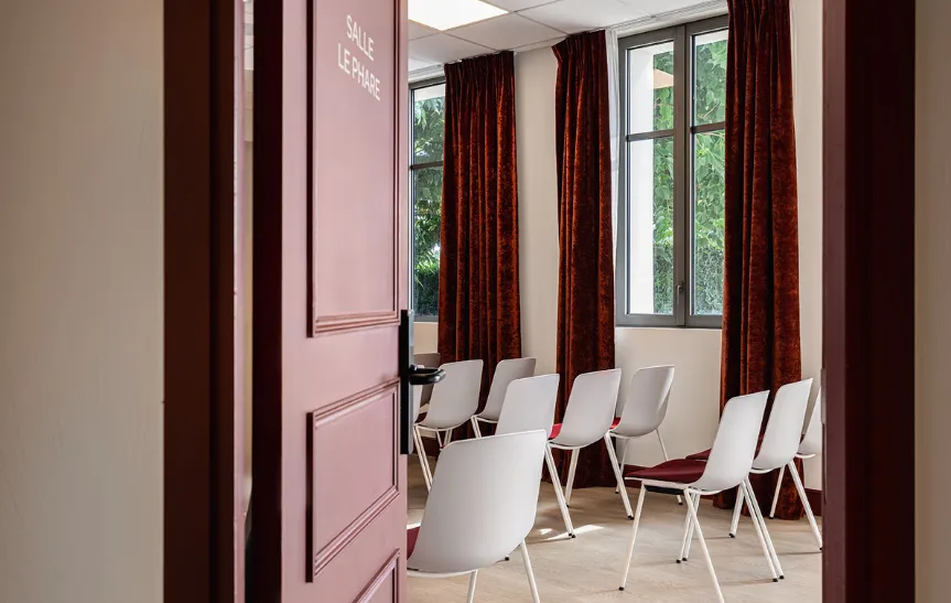 Vea a través de una puerta burdeos entreabierta con la inscripción «SALLE LE PHARE», que da a una sala de reuniones dispuesta en hileras de sillas blancas y rojas. La sala está iluminada por ventanas con cortinas de terciopelo burdeos.