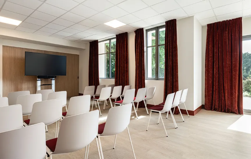 Luminosa sala de reuniones amueblada en estilo teatro con numerosas sillas blancas y rojas, una gran pantalla de televisión montada en la pared y grandes ventanales revestidos con cortinas de terciopelo burdeos