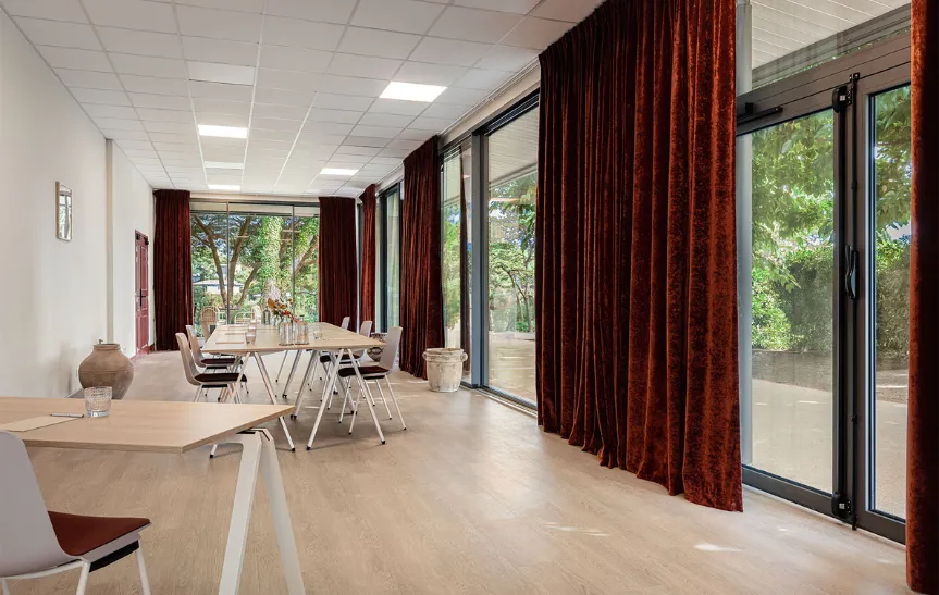 Sala de reuniones grande y luminosa con grandes ventanales con vistas a la vegetación, enmarcada por gruesas cortinas de terciopelo burdeos. En el suelo de madera clara hay una larga mesa de reuniones y mesas de trabajo individuales.