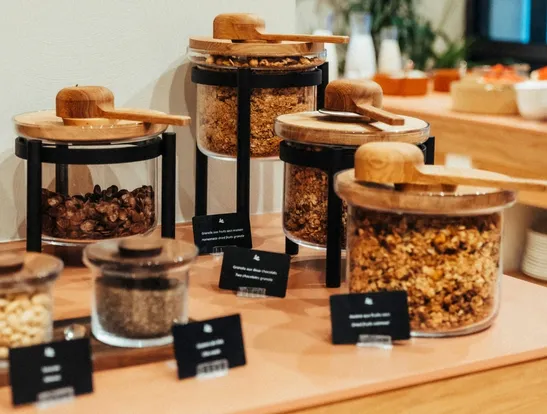 An assortment of cereals, muesli and nuts in glass jars with wooden lids and spoons. Each jar is clearly labelled on the buffet.