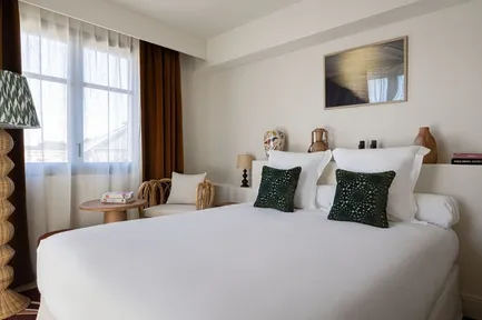 Chambre d'hôtel lumineuse avec un grand lit blanc, des coussins vert émeraude, une fenêtre avec rideaux blancs et bordeaux, et une lampe sur pied en osier