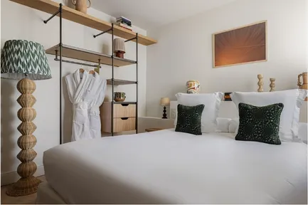 Gros plan sur un grand lit d'hôtel avec des coussins vert foncé. La pièce s'ouvre directement sur un balcon avec vue sur la verdure grâce à une porte-fenêtre. Une grande lampe sur pied en rotin est visible sur le côté droit.