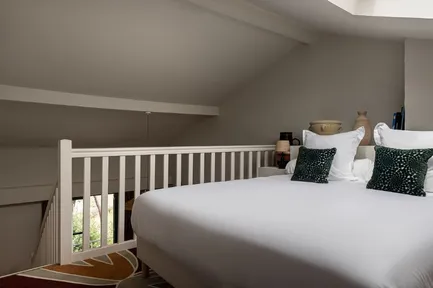 Lit double blanc sur une mezzanine mansardée avec une balustrade blanche. Coussins décoratifs vert foncé sur fond de murs gris clair.