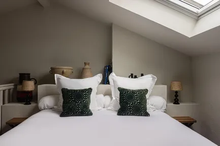 Gros plan sur un grand lit double d'hôtel avec une literie blanche et deux coussins décoratifs vert foncé. La tête de lit est décorée de petites lampes de chevet et de divers objets décoratifs. Une étagère supérieure est visible avec quelques livres