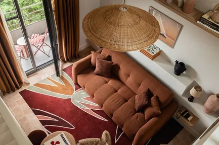 Vue plongeante sur un salon au style bohème et chaleureux, avec un canapé couleur terre cuite, un grand tapis bordeaux à motifs floraux, et une suspension circulaire en osier. Une porte-fenêtre donne sur un balcon