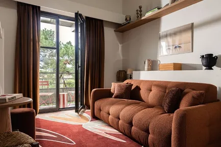 Salon d'hôtel moderne et chaleureux avec un canapé couleur terre cuite et deux fauteuils en rotin. La pièce est décorée d'un grand tapis à motifs floraux rouge et d'une suspension en paille tressée