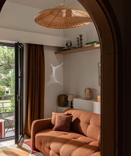 Salon d'hôtel moderne et chaleureux avec un canapé couleur terre cuite et deux fauteuils en rotin. La pièce est décorée d'un grand tapis à motifs floraux rouge et d'une suspension en paille tressée