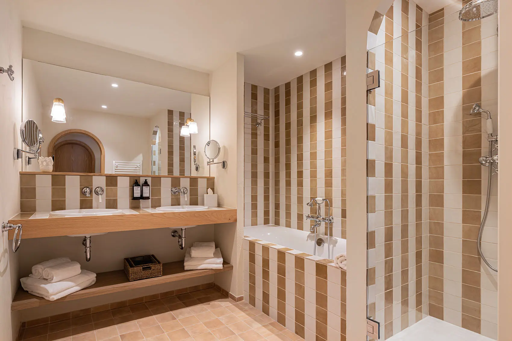 Amplio baño de hotel con dos lavabos y una ventana grande, renovado con una pieza adicional de madera. El baño y la ducha están cubiertos con azulejos verticales a rayas blancas y beige, con pisos de terracota.