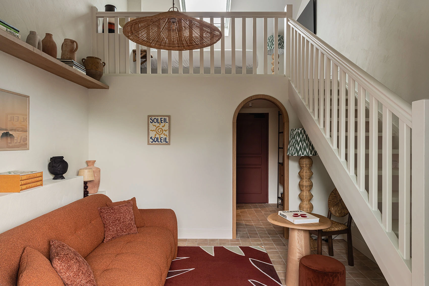 Interior de una suite dúplex con una escalera que conduce a un entresuelo y un sofá de terracota en primer plano. El espacio también incluye un arco que conduce a otra habitación, una alfombra roja estampada y decoraciones de pared y jarrones en las estanterías