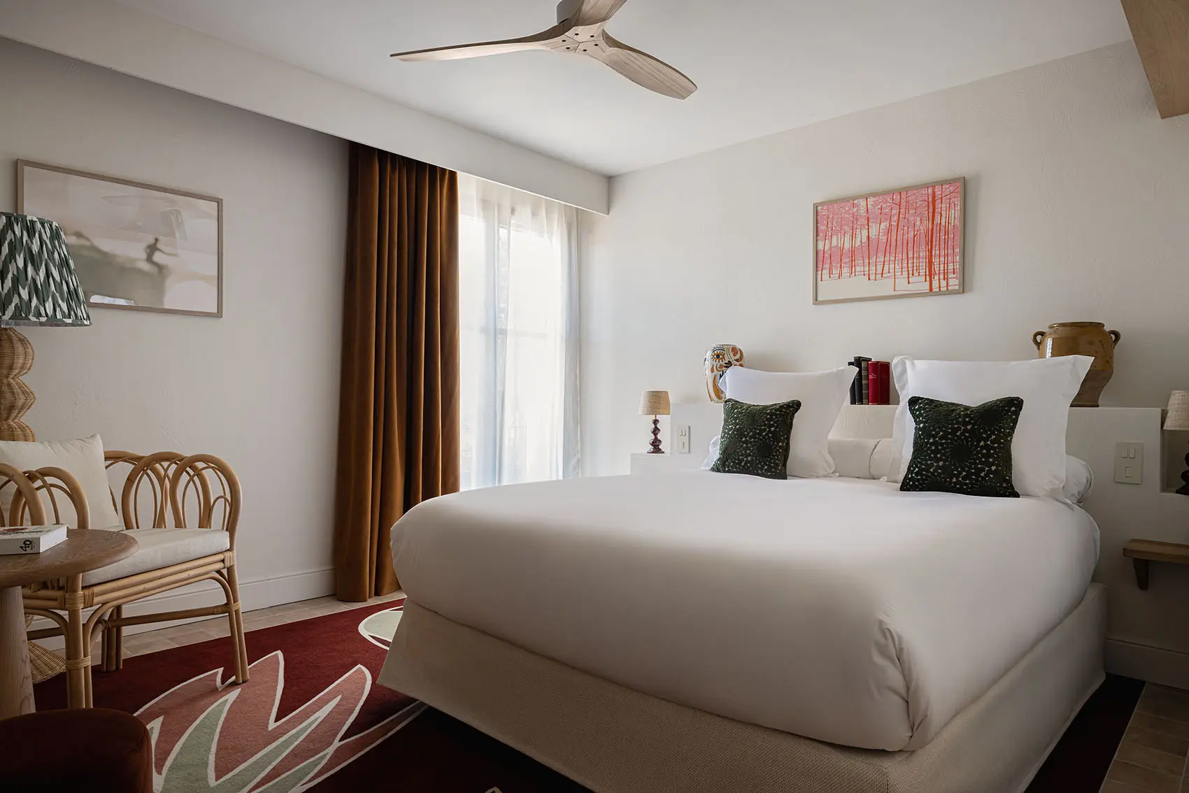 Habitación de hotel moderna y luminosa con un amplio dormitorio con ropa blanca, rincones decorativos de color verde oscuro y un ventilador tecnológico. Junto a la cama, hay un sillón de ratán y una mesita auxiliar pequeña sobre una alfombra roja texturizada