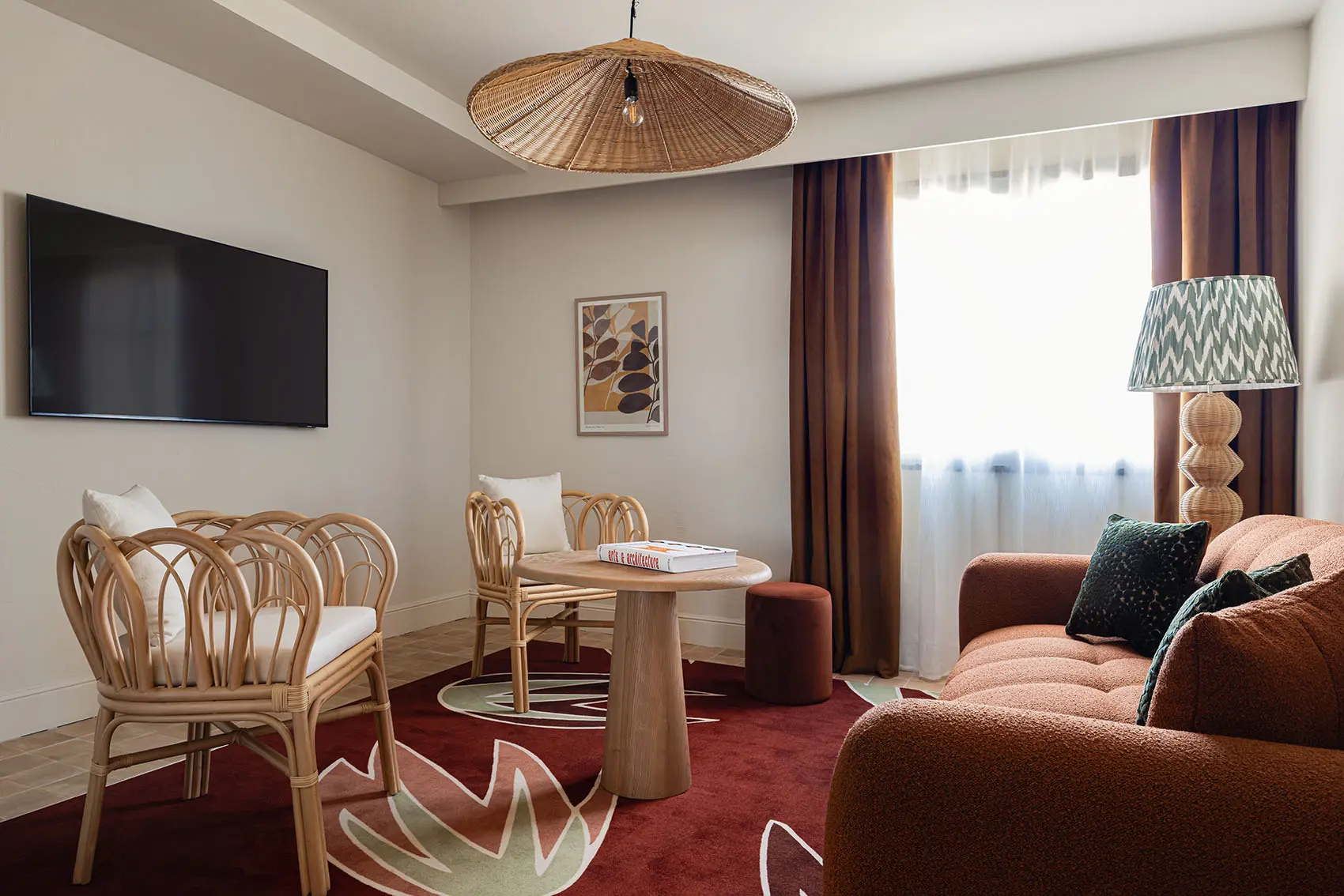 El salón del hotel es moderno y cálido, con tonos terracota y elementos naturales. La habitación incluye un sofá texturizado, dos surcos de papel y un gran televisor montado en la pared, y está iluminada por una lámpara intermitente y una lámpara de pie de fibra natural.