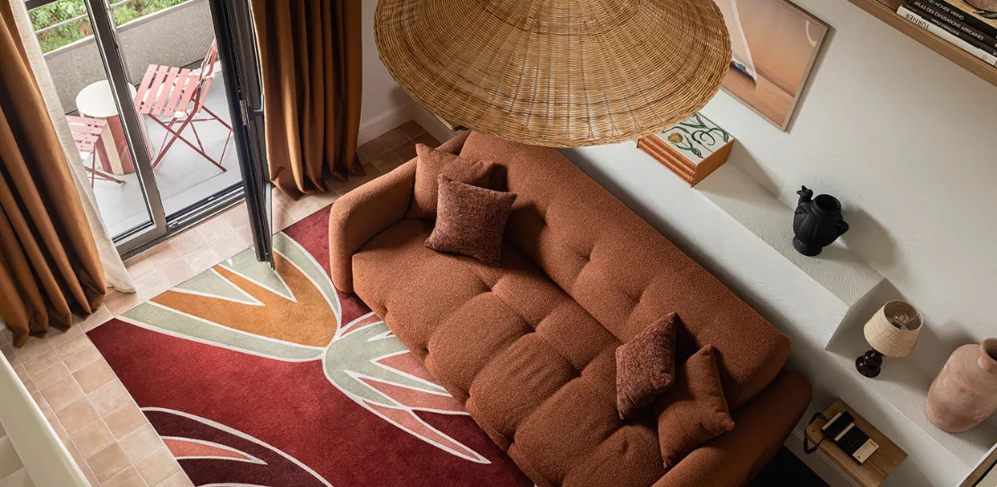 Vista aérea de la sala de estar de una suite de hotel: un gran sofá de color terracota se coloca sobre una alfombra estampada en rojo y verde. Arriba, una gran suspensión de paja tejida. A la izquierda, una puerta de cristal conduce a un pequeño balcón exterior