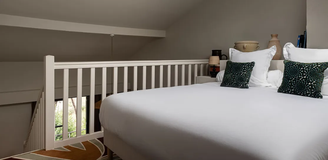 Primer plano en una cama grande con ropa de cama blanca en una habitación abuhardillada o en el entresuelo. La cabecera está decorada con cojines y jarrones de color verde oscuro. Una balaustrada blanca da a la planta inferior y en primer plano se ve una alfombra estampada