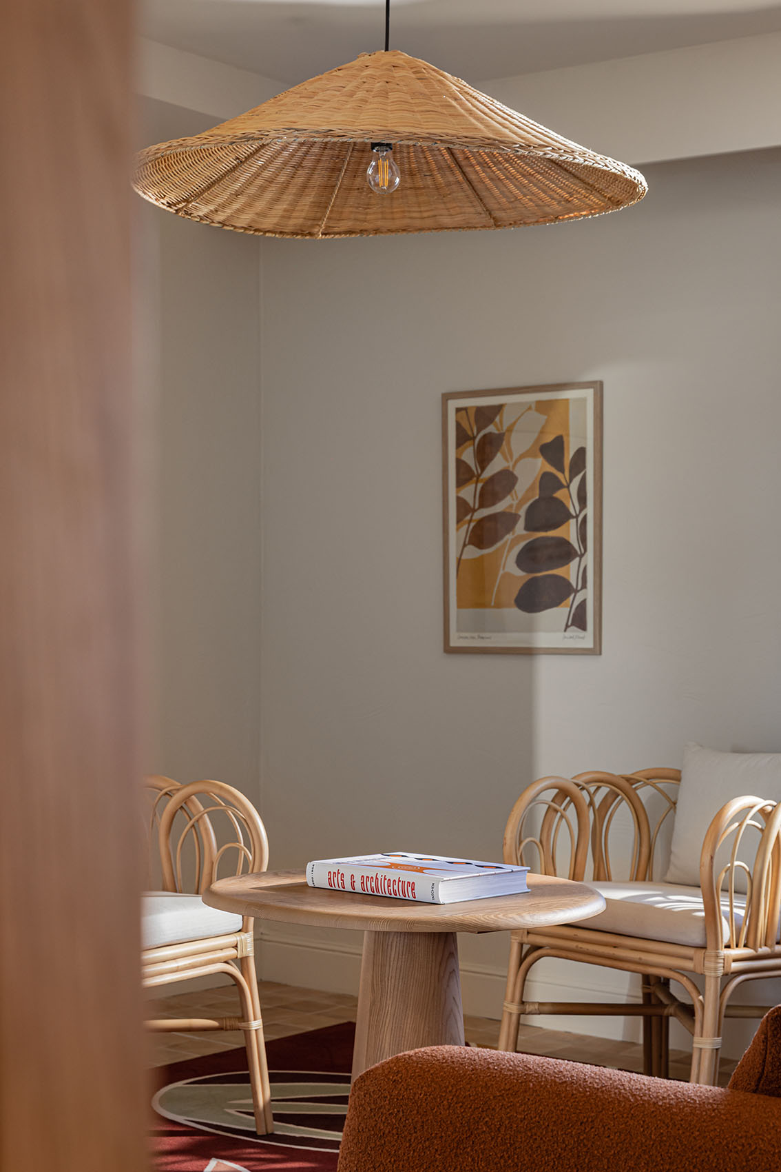 Habitación de hotel moderna y luminosa con una cama grande de lino blanco, cojines decorativos de color verde oscuro y un ventilador de techo. Junto a la cama, hay un sillón de ratán y una mesita auxiliar pequeña sobre una alfombra roja texturizada