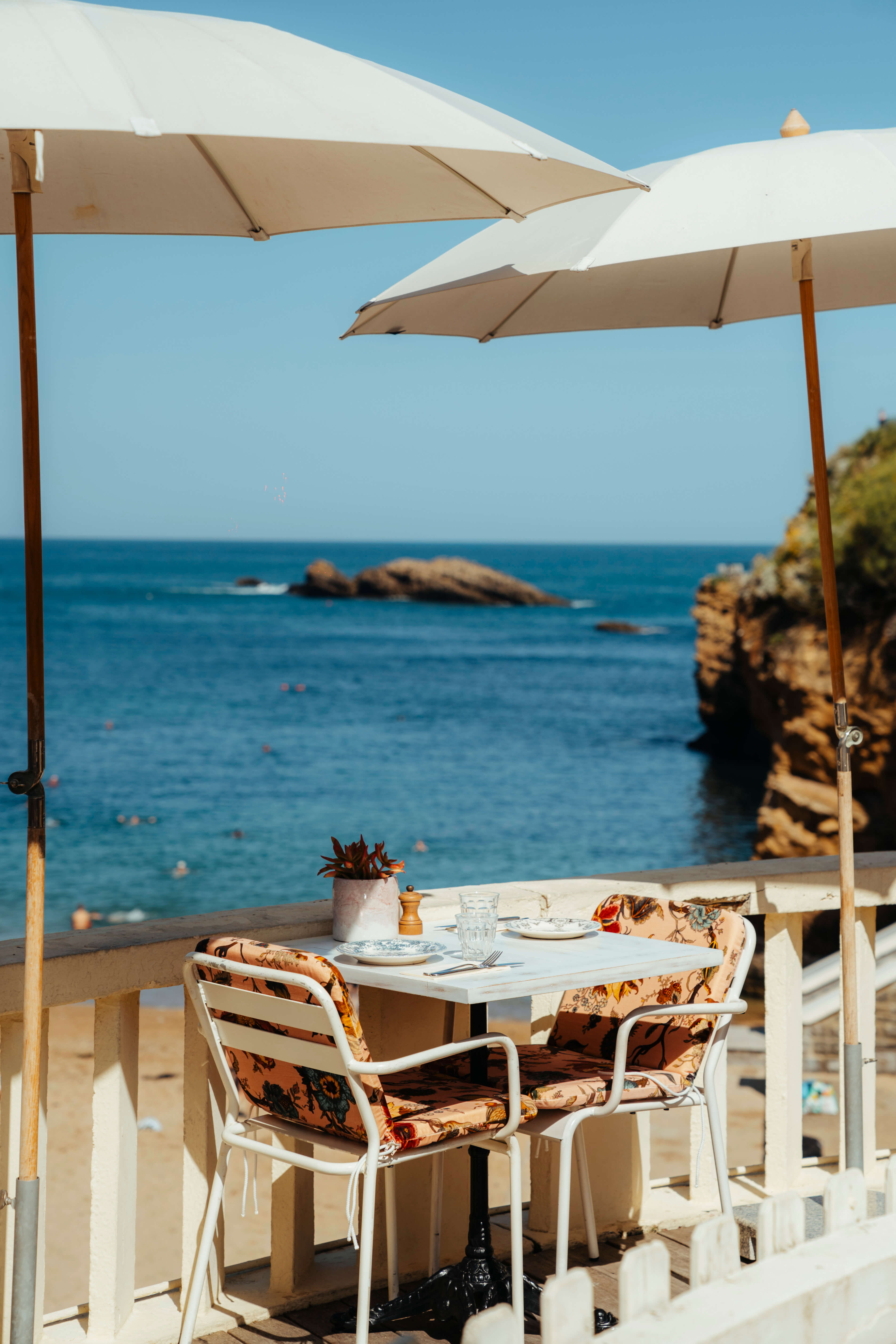 Table de café en plein air pour deux, sous des parasols blancs, avec vue sur la mer bleue ensoleillée, des rochers et des personnes nageant au loin.