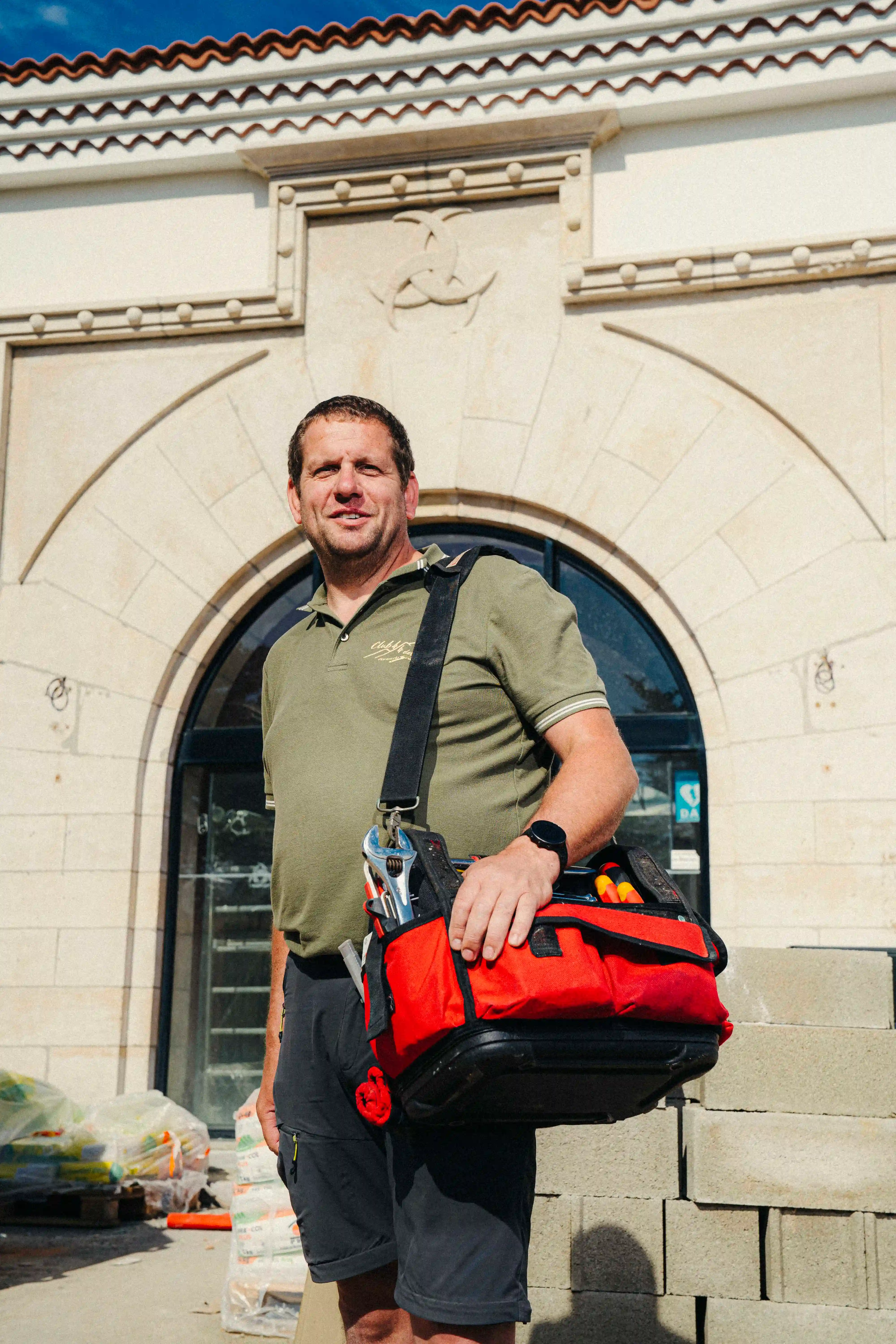 homme d'affaires souriant portant un polo et un sac à outils professionnel rouge, debout devant une façade en pierre avec une arche.