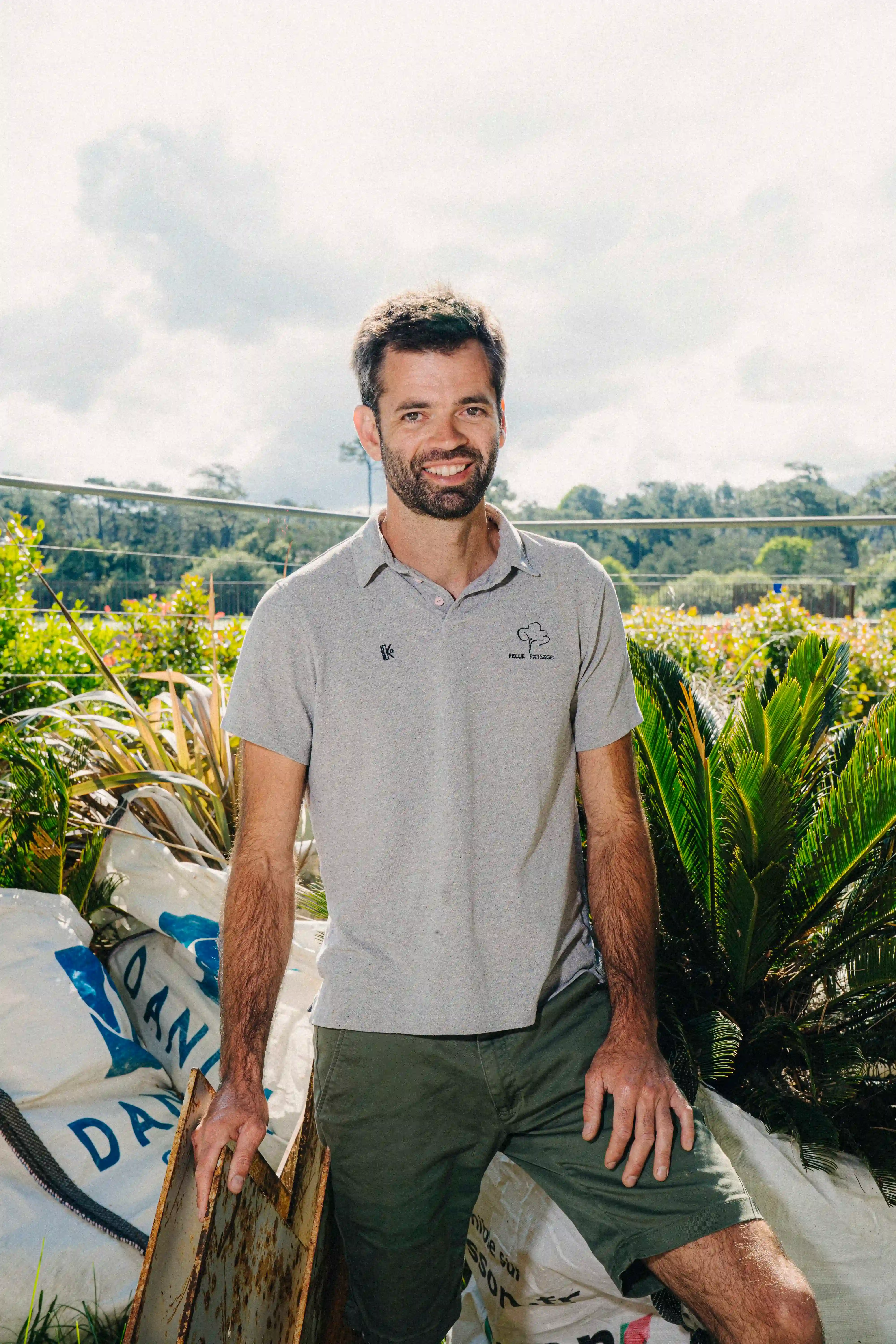 Homme souriant portant un polo gris et un pantalon kaki, se tenant debout au milieu de plantes de pépinière et de sacs d'engrais. Le ciel est partiellement nuageux et on aperçoit des arbres et des clôtures en arrière-plan.