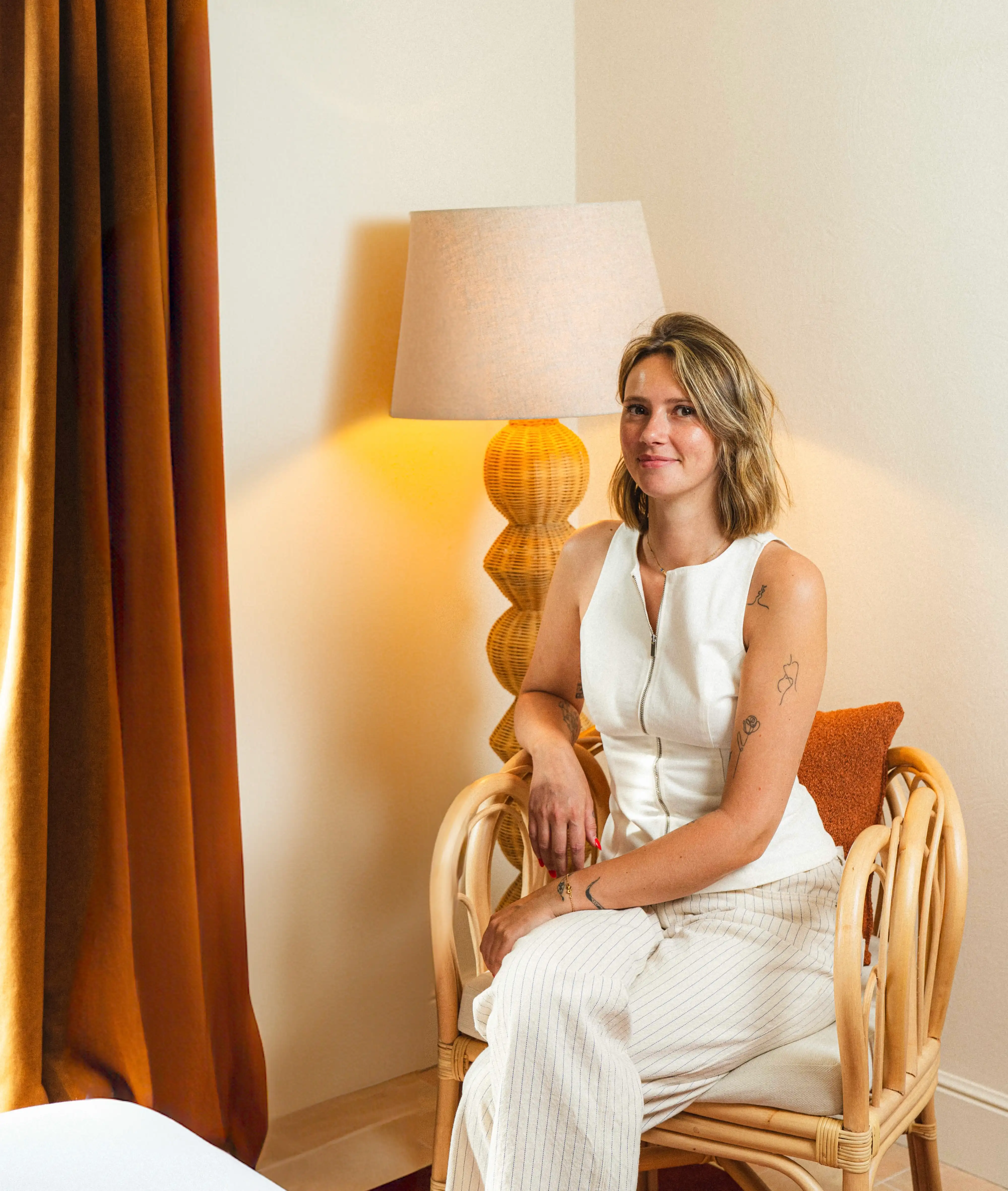 Femme souriante vêtue d'un haut blanc sans manches et d'un pantalon rayé clair, assise dans un fauteuil en rotin devant une lampe à abat-jour et un rideau couleur rouille.
