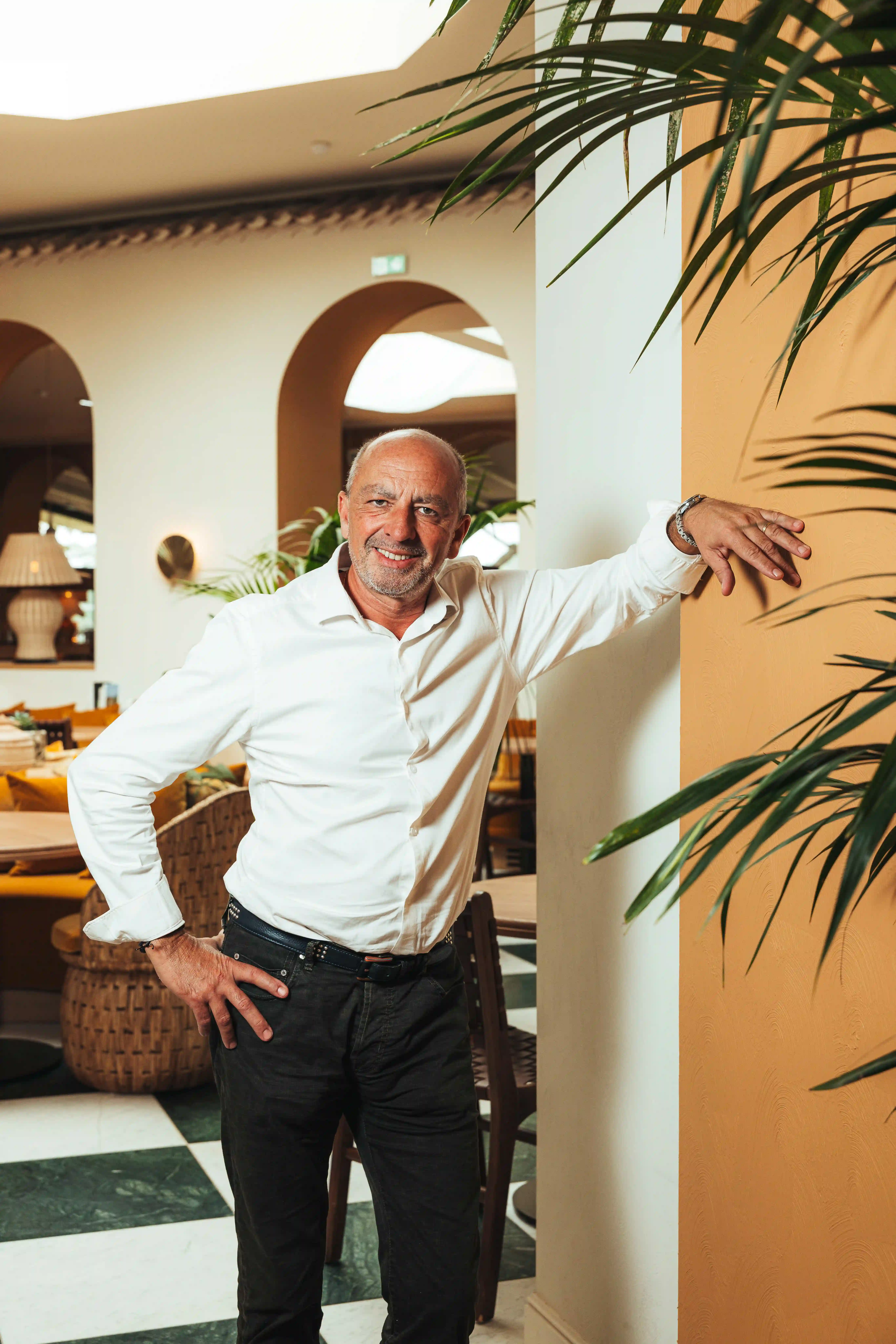 Homme souriant portant une chemise blanche et un pantalon noir, se tenant debout dans le patio au milieu de plantes.