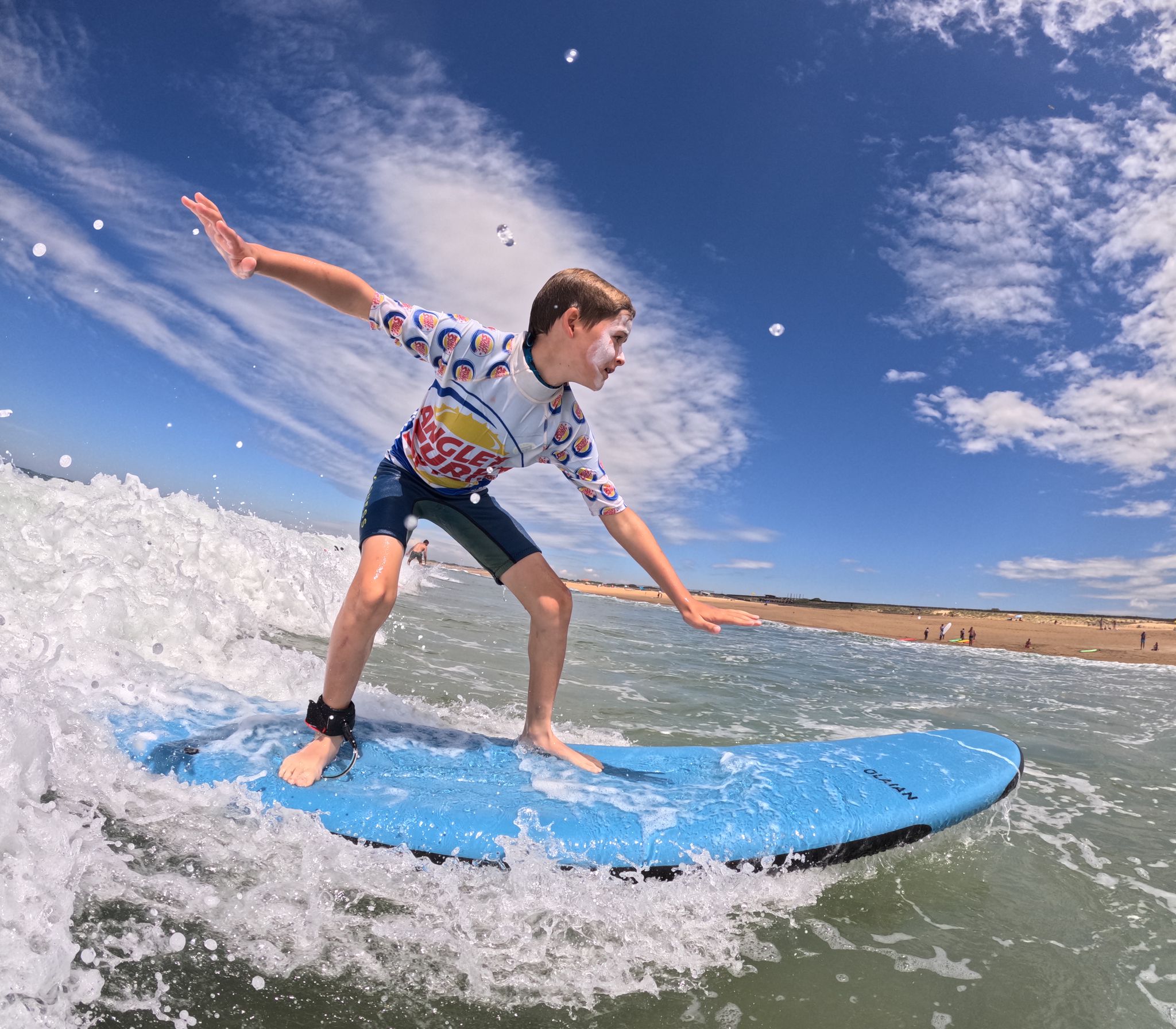 Un homme et une femme portant des combinaisons de plongée sur des planches de surf, attendant une vague dans l'océan. La femme est allongée sur sa planche tandis que l'homme est en train de ramer. Le ciel est bleu et dégagé