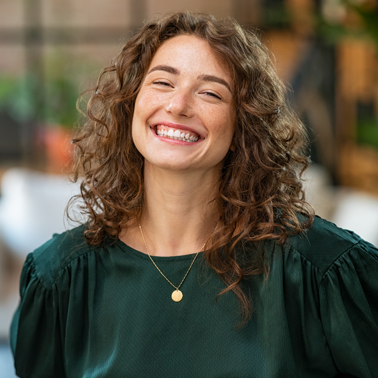 Smiling woman with curly brown hair wearing a green blouse and gold necklace with a round pendant.