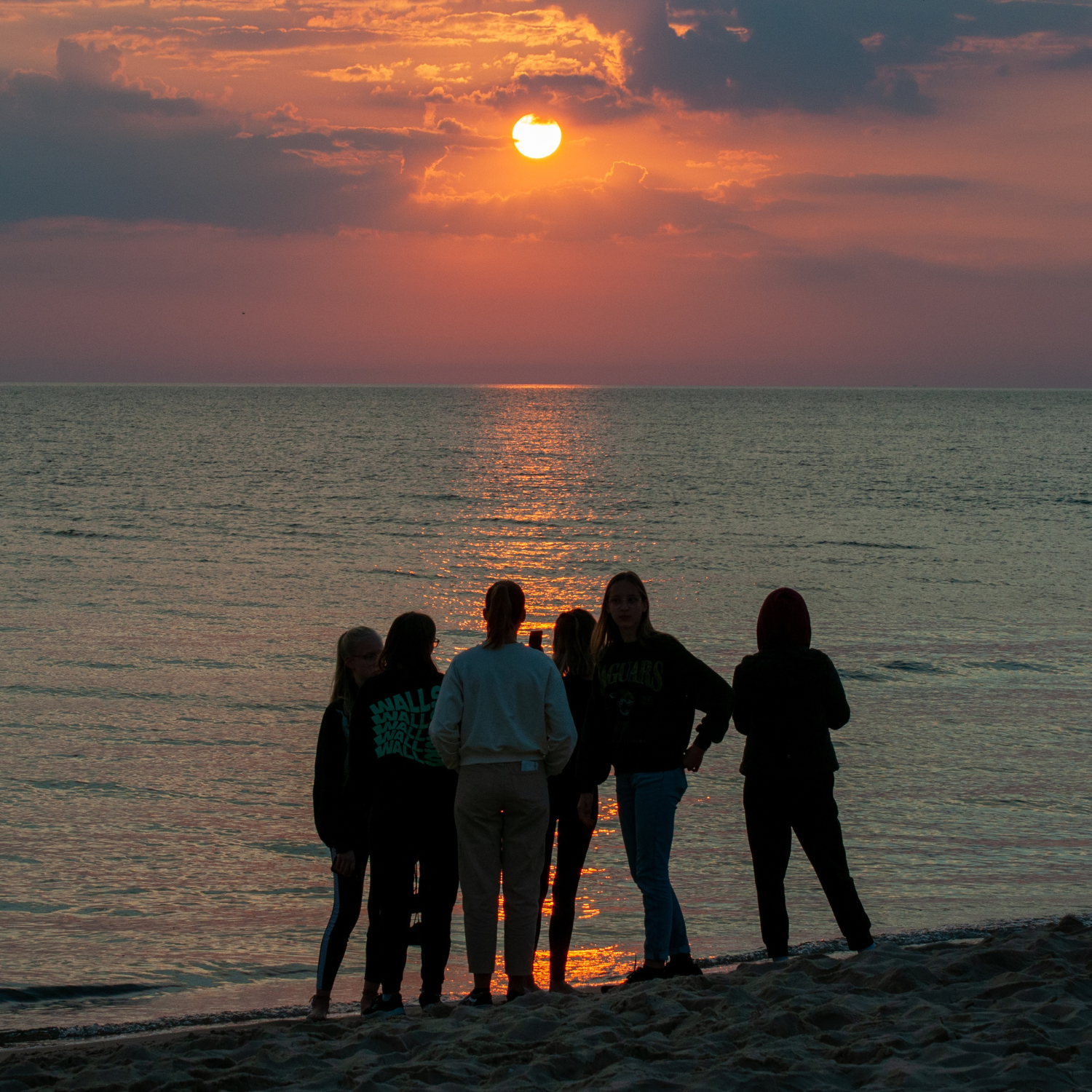 Group of six people standing on a sandy beach watching the sun setting over the calm ocean.