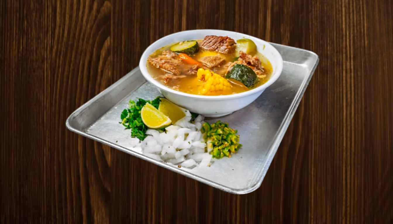 Bowl of beef soup with corn, zucchini, and carrot served on a metal tray with lime wedges, chopped onions, cilantro, and sliced green chilies.