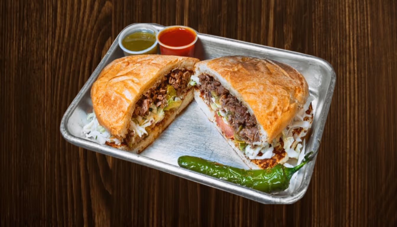 Mexican torta sandwich cut in half on a metal tray with shredded cabbage, meat, tomato, pickled jalapeños, grilled cheese, a green chili pepper, and two small cups of green and red sauce.