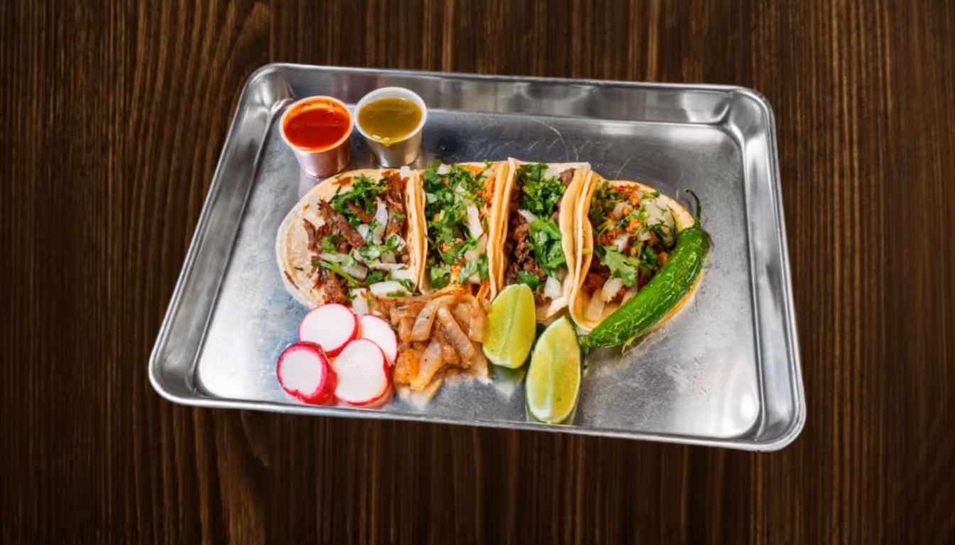 Metal tray with four tacos topped with cilantro and onions, lime wedges, grilled onion, radish slices, a grilled green chili, and two small containers of red and green salsa.