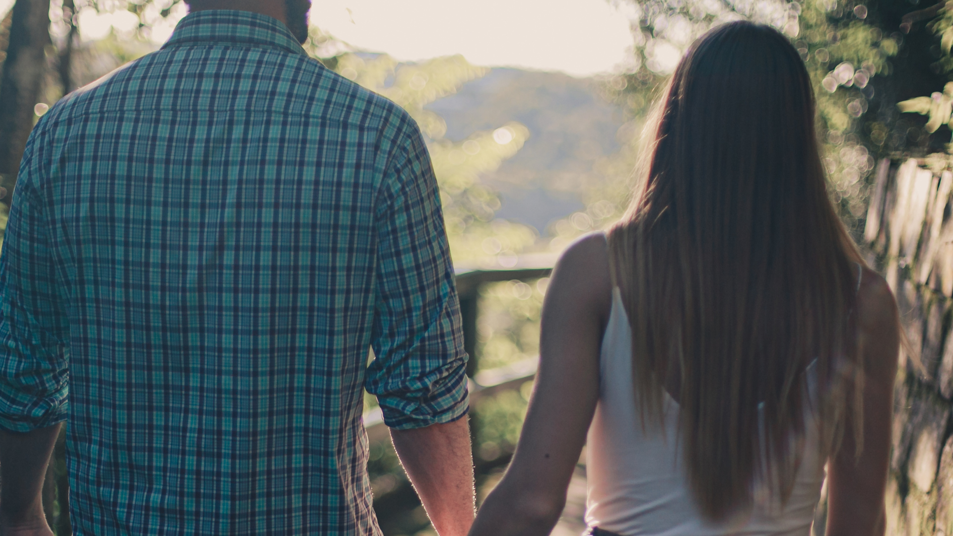 Couple holding hands walking outdoors with blurred trees and sunlight in the background.