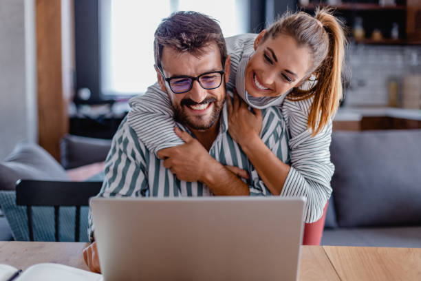 Smiling young woman hugging a man from behind as they look at a laptop together in a cozy living room.