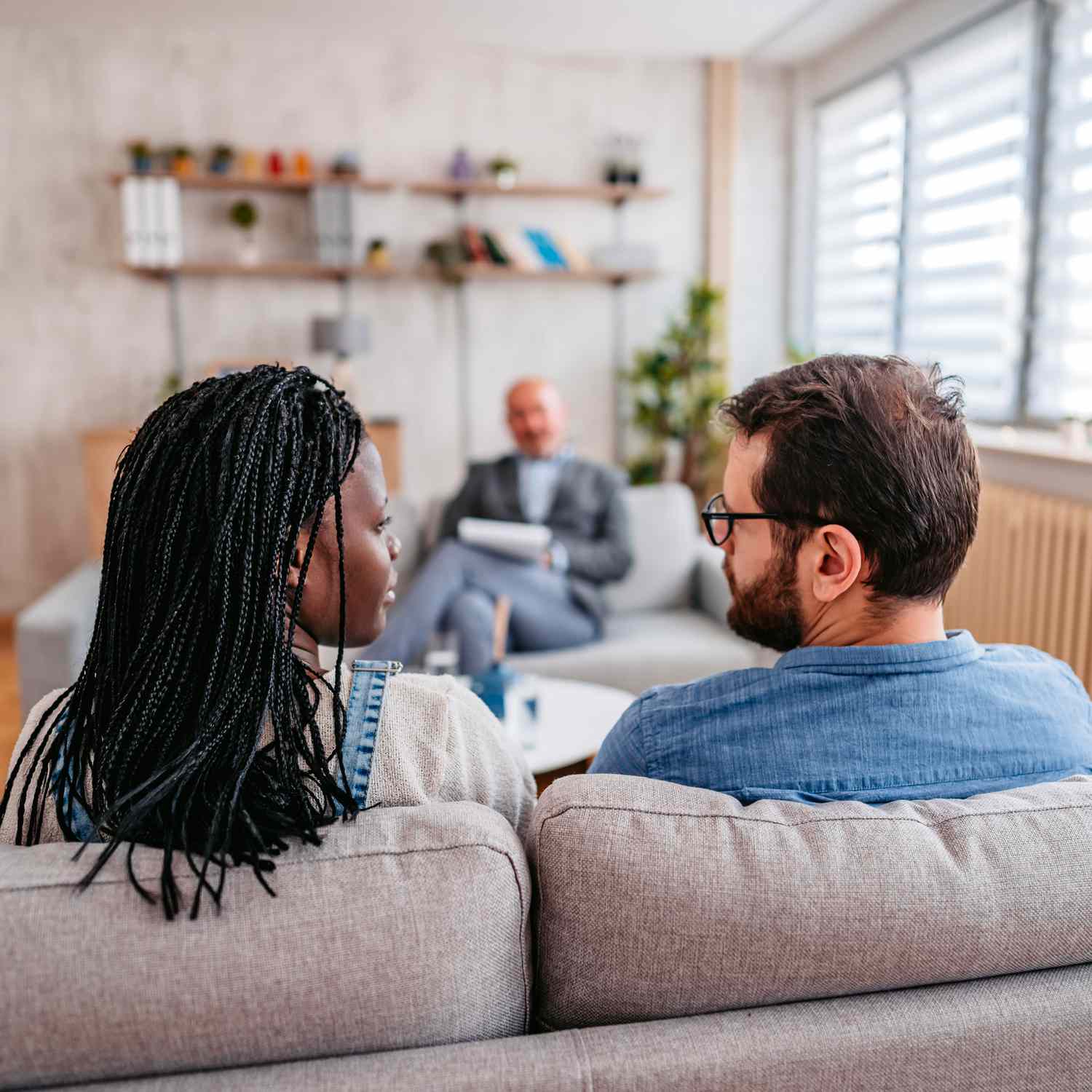 A couple that is sitting on a couch in a room facing a therapist. You can see the back of their heads and the therapist who is facing them.  