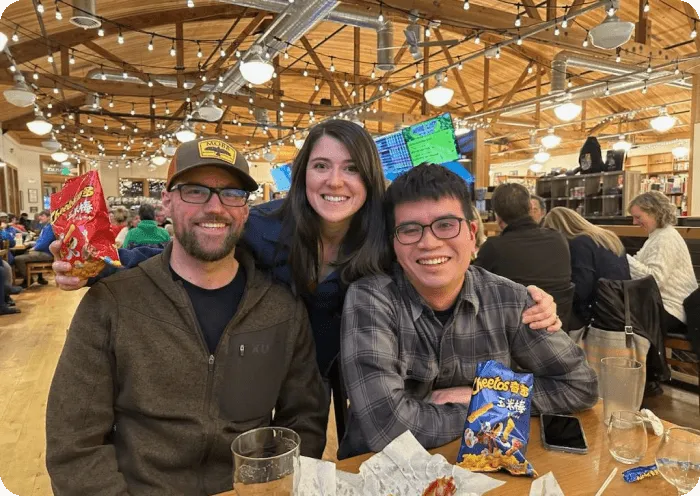 Three friends smiling and posing at a wooden table in a warmly lit restaurant, with two holding bags of snacks.