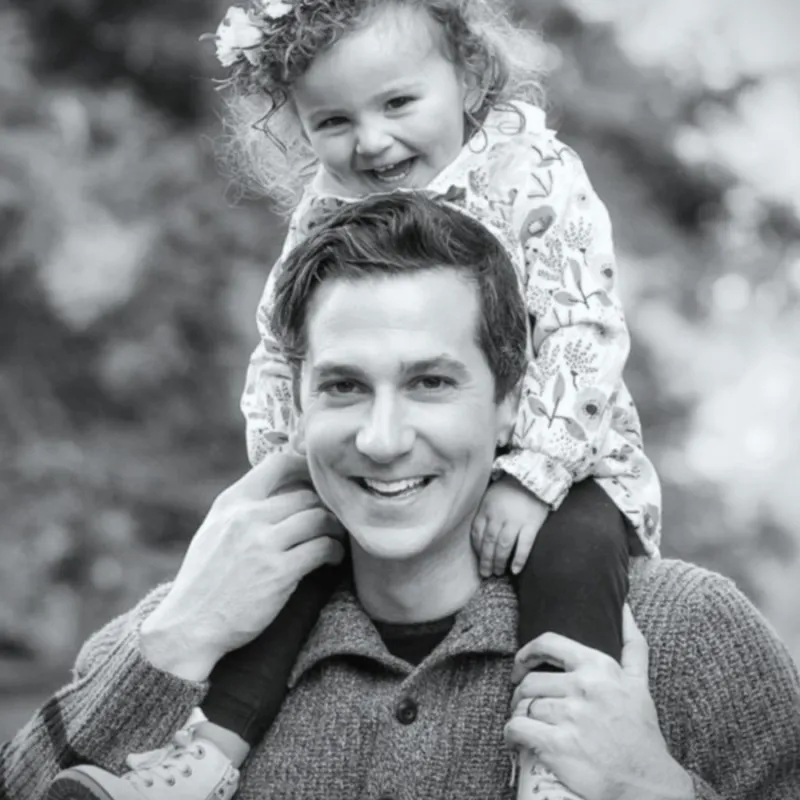 Smiling man carrying a happy toddler girl on his shoulders outdoors.