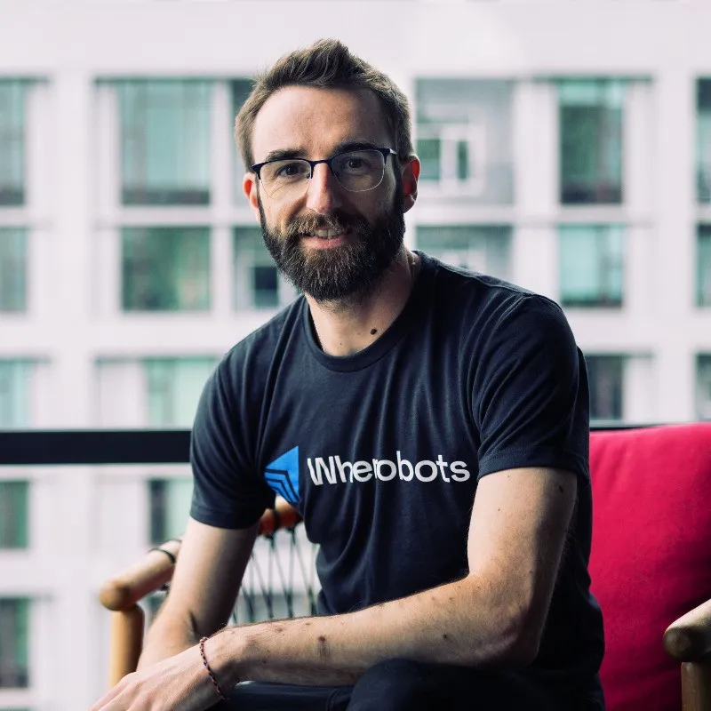 Man with glasses and beard wearing a black Wherebots t-shirt sitting on a chair with a blurred building background.