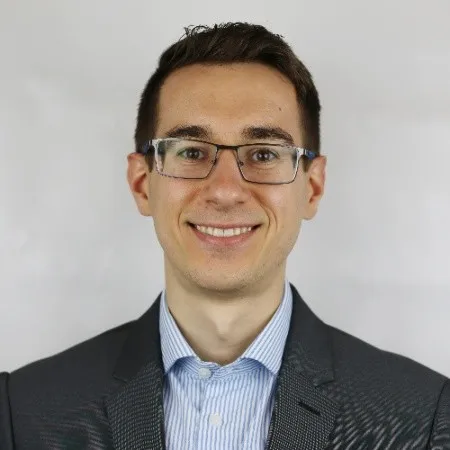 Smiling man wearing glasses, a striped dress shirt, and a dark blazer against a plain light background.