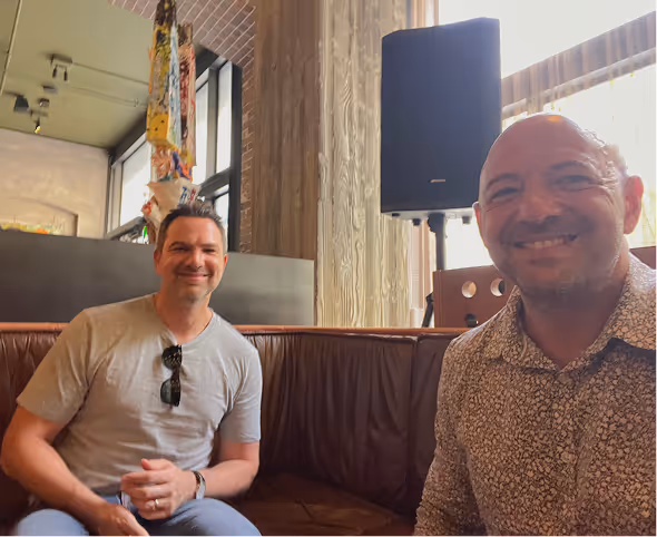 Two men smiling while sitting on a brown leather couch in a room with large windows and wooden walls.