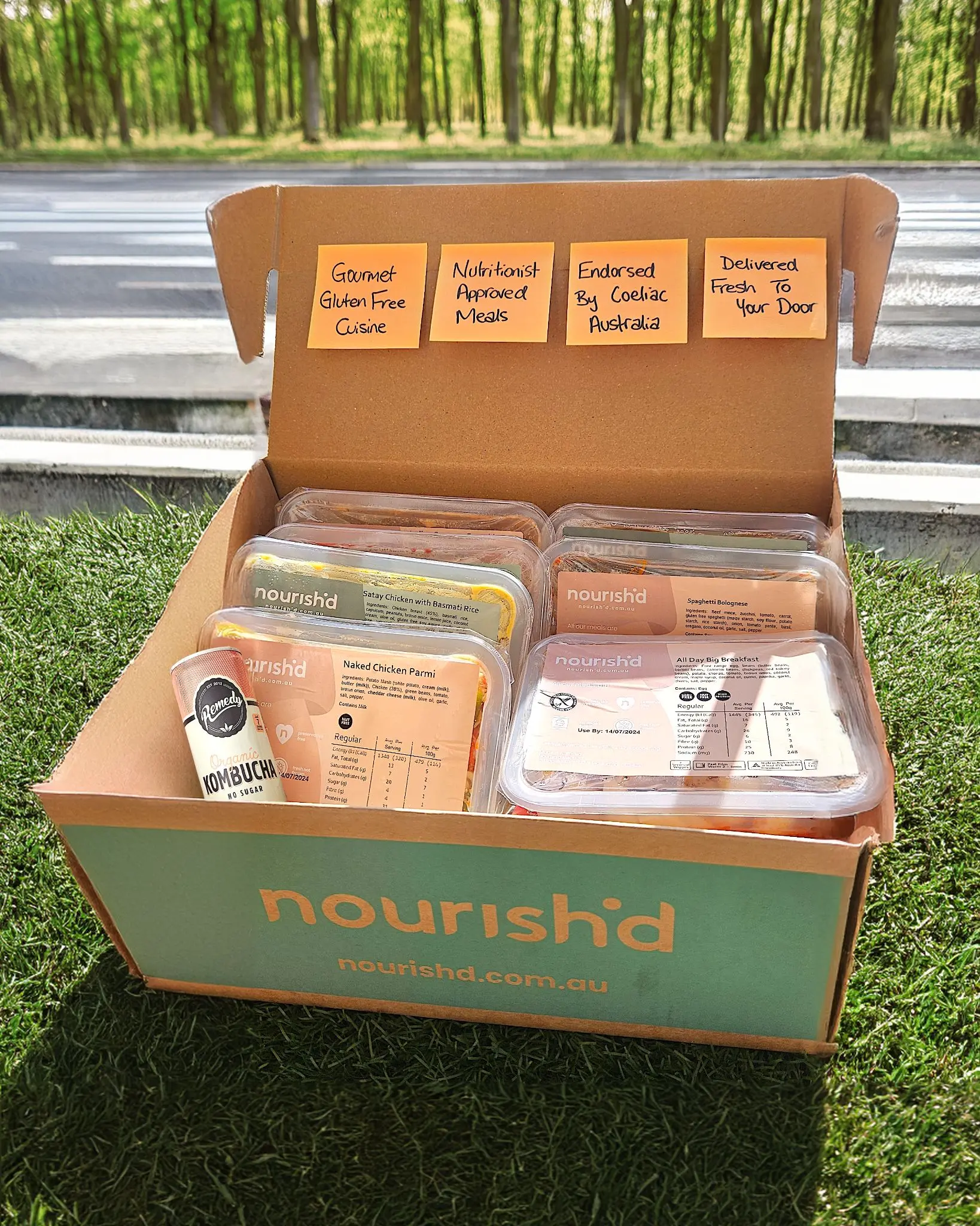 Open Nourishd meal delivery box with four sticky notes reading Gourmet Gluten Free Cuisine, Nutritionist Approved Meals, Endorsed By Coeliac Australia, Delivered Fresh To Your Door, containing prepackaged meals and a can of Remedy Organic Kombucha.